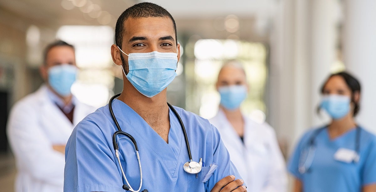 Confident multiethnic male nurse in front of his medical team looking at camera wearing face mask
